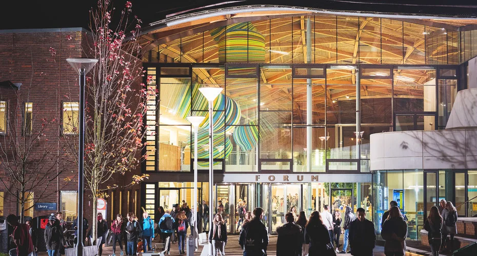 People walking in front of the Forum building at the university of exeter, illuminated at night.